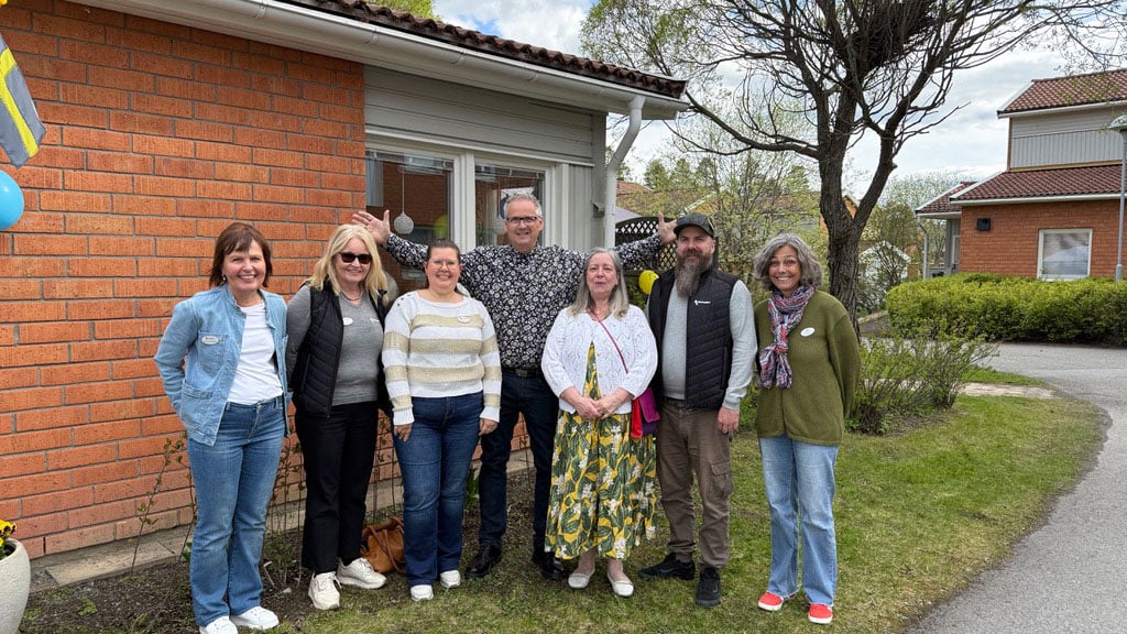 Grupp av människor utanför ett tegelhus, med träd och gräsmatta i bakgrunden