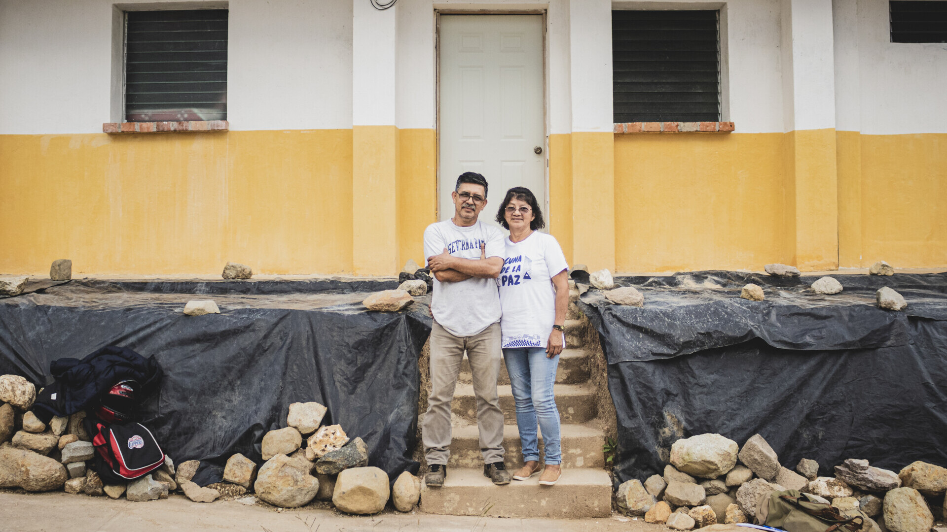 Man och kvinna står framför en byggnad med gul och vit fasad, omgivna av stenar och svarta plastskynken