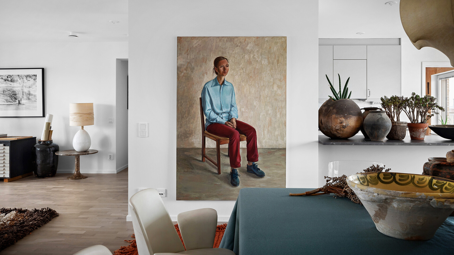 Interiör av ett vardagsrum med ett stort porträtt av en sittande person i blå skjorta och röda byxor. Rummet har vita väggar, en bordslampa, och flera krukväxter på en hylla
