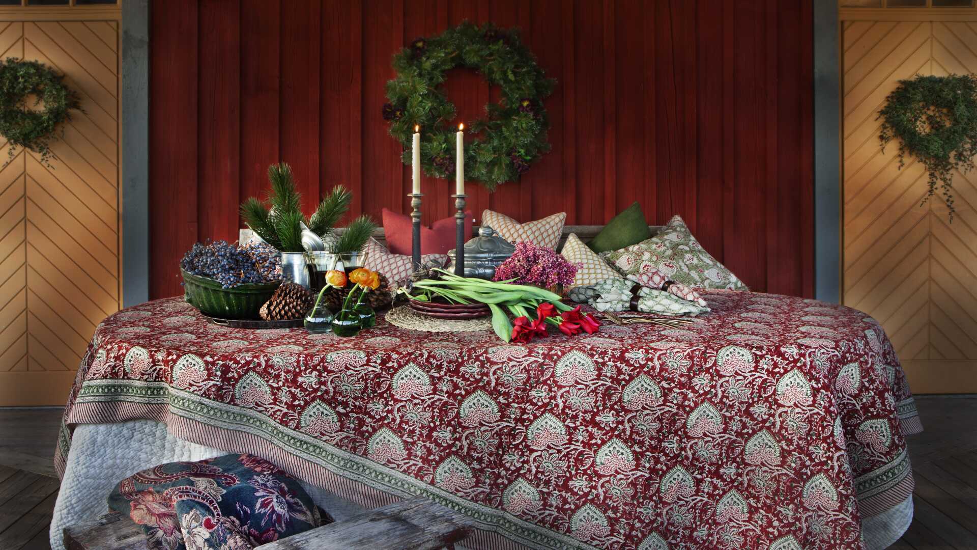 Dukat bord med röd duk och blommor samt ljusstakar, mot en mörkröd trävägg med kransar