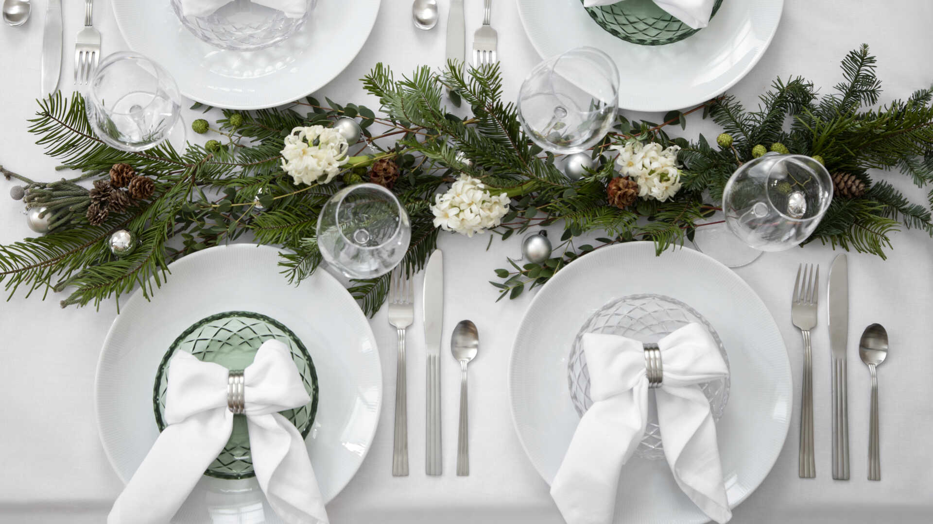Festligt dukat bord med vita tallrikar, gröna servetter knutna med vita rosetter, och dekoration av granris och vita blommor