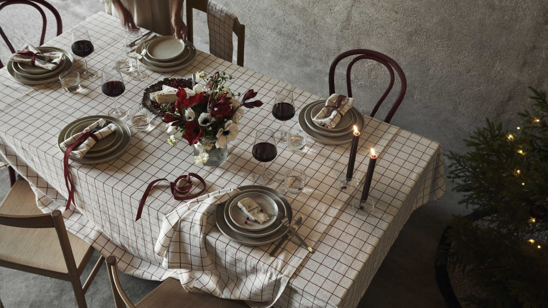 Dukat matbord med rutigt bordduk, tallrikar, bestick, vinglas och blomsterarrangemang, omgiven av trästolar och en julgran