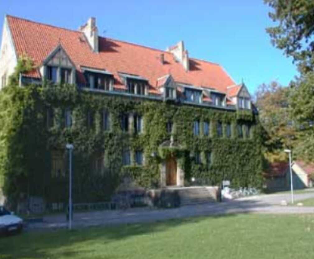 Stenbyggnad med rött tegeltak täckt av murgröna och omgiven av grönska