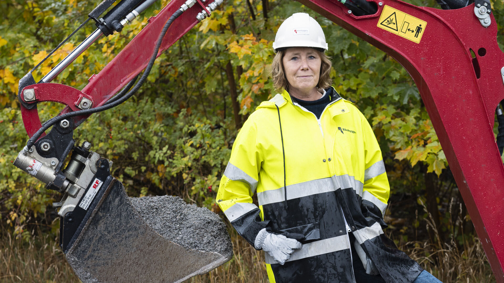 Kvinna i varselkläder och hjälm står vid en grävmaskin framför höstfärgade träd