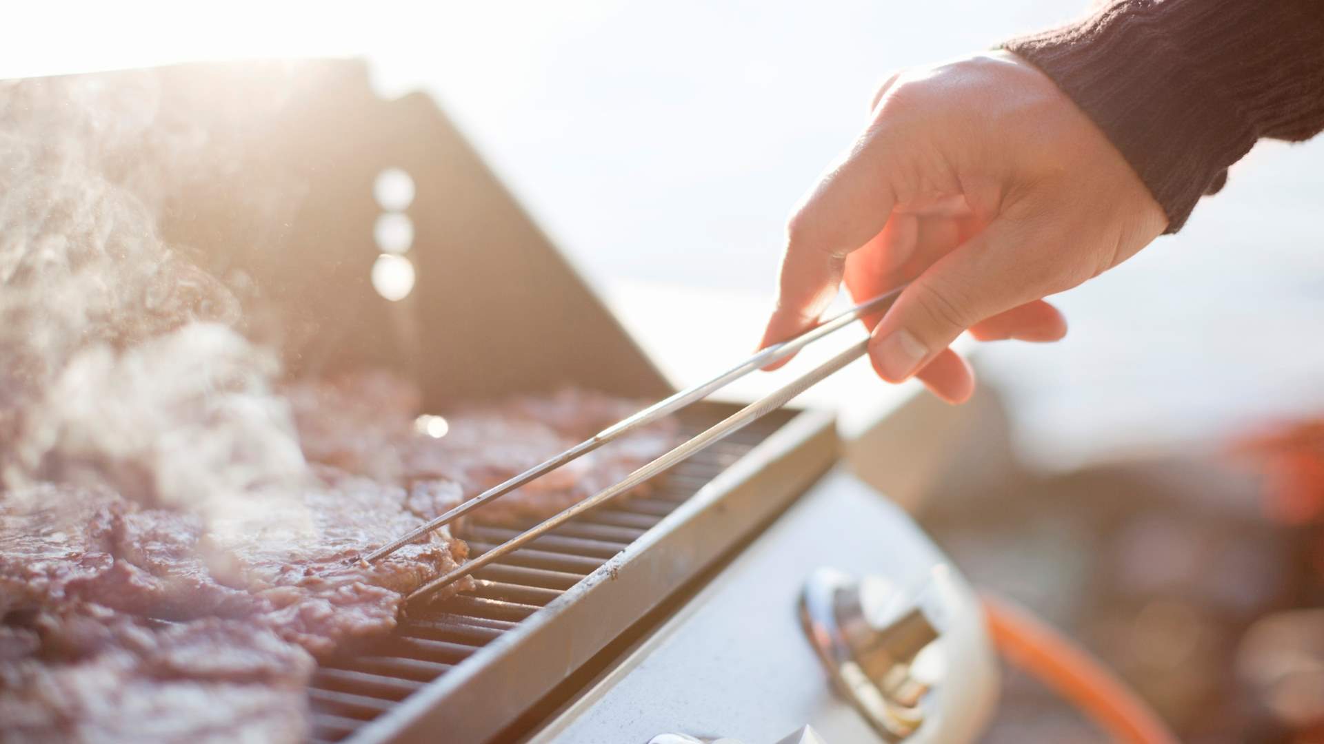 Hand som använder grilltång för att tillaga kött på en utomhusgrill med solens strålar i bakgrunden