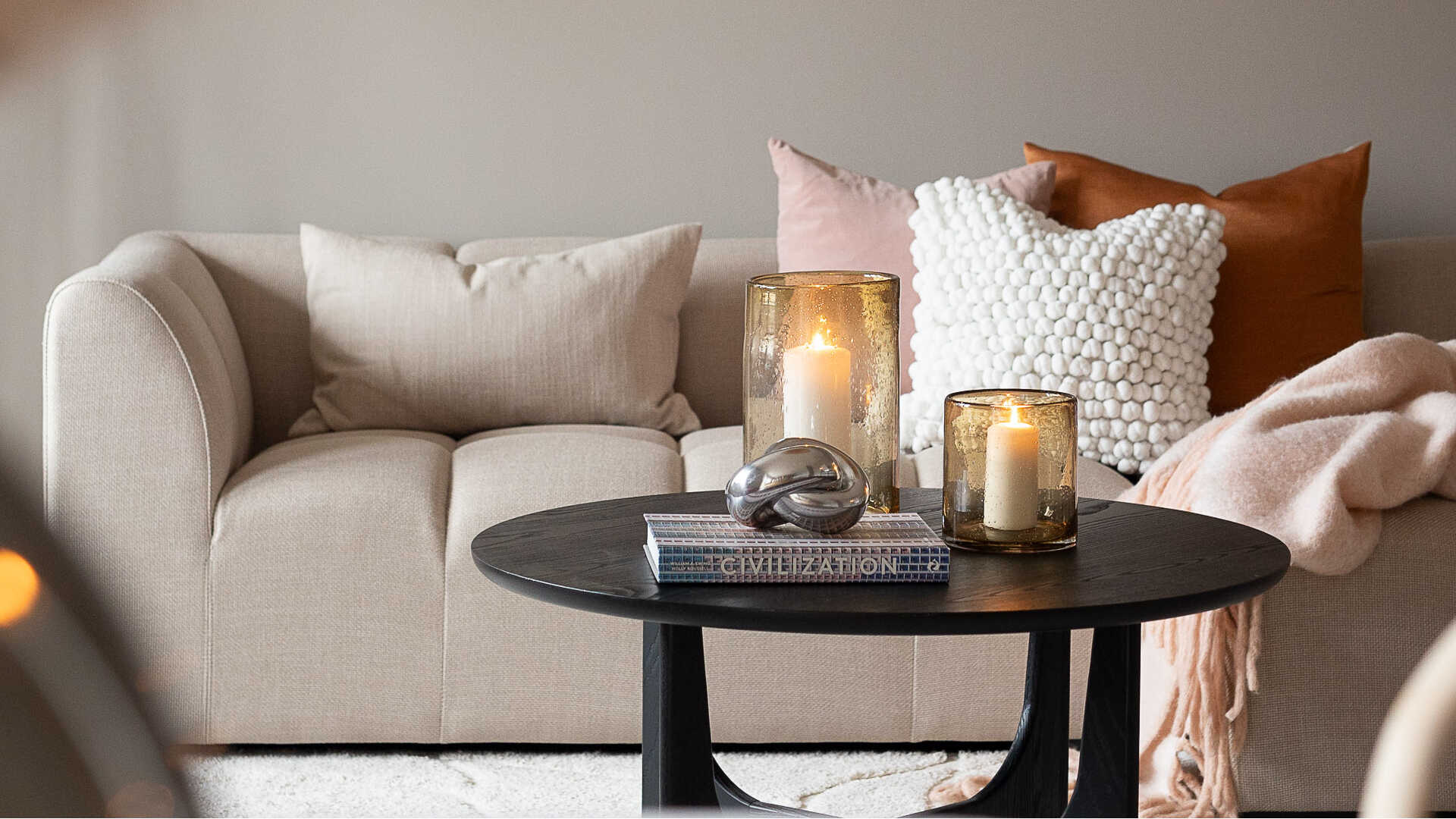 Soffgrupp med beige och rosa kuddar, svart soffbord med tända ljus och en bok i vardagsrumsmiljö