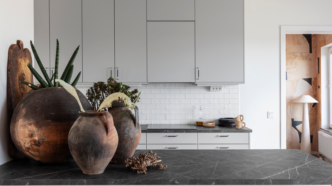 Modernt kök med grå skåp och svarta bänkskivor, dekorerat med stora rustika lerkrukor och växter. Vitt kakel i bakgrunden och en öppen dörr som leder mot ett mönstrat rum