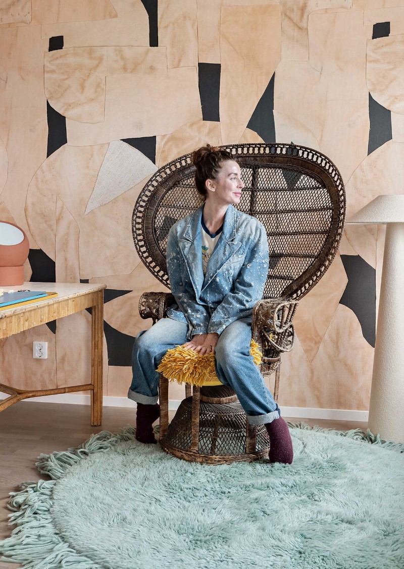 Marie Olsson Nylander i jeansjacka och jeans sitter på en rottingstol i ett rum med ljusblå matta och mönstrad vägg