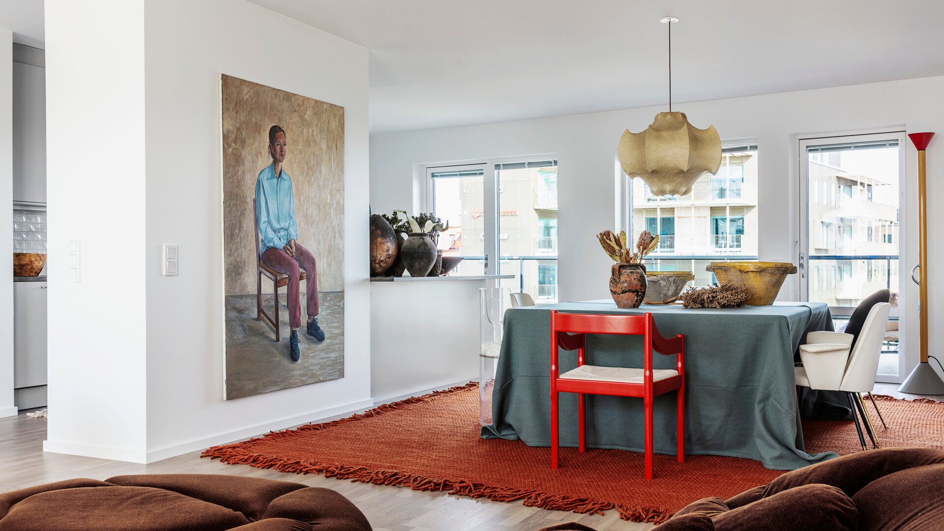 Modernt vardagsrum med en stor målning av en person på en stol, ett matbord med en röd stol och olika dekorativa föremål, samt stora fönster i bakgrunden