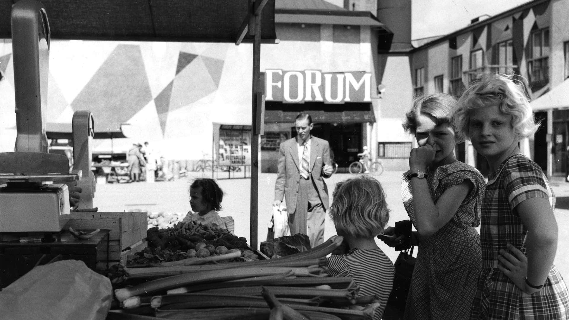 Marknadsstånd med grönsaker framför en byggnad med skylten 