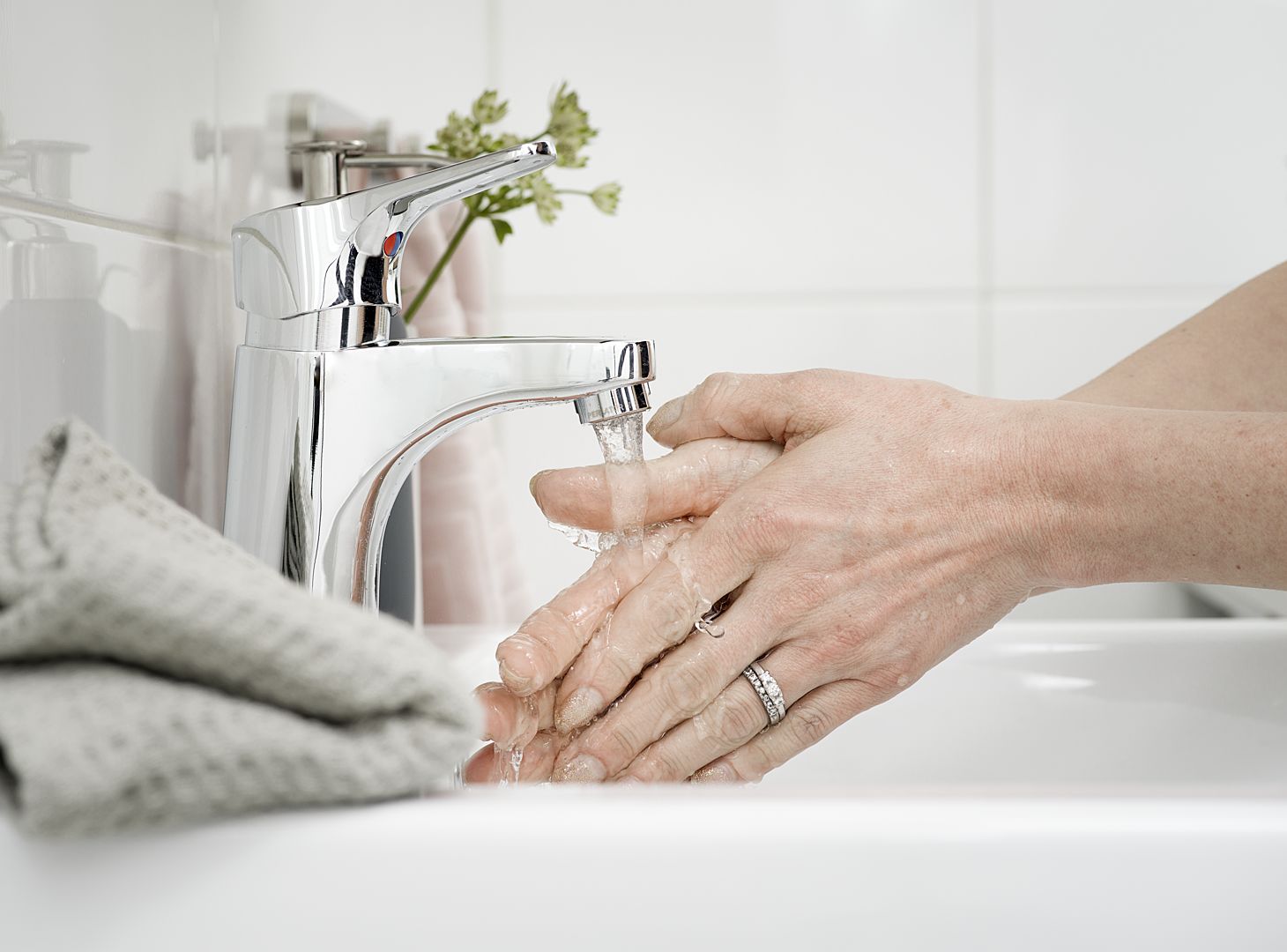 Händer tvättas under rinnande vatten från en silverfärgad kran, med en grå handduk i förgrunden
