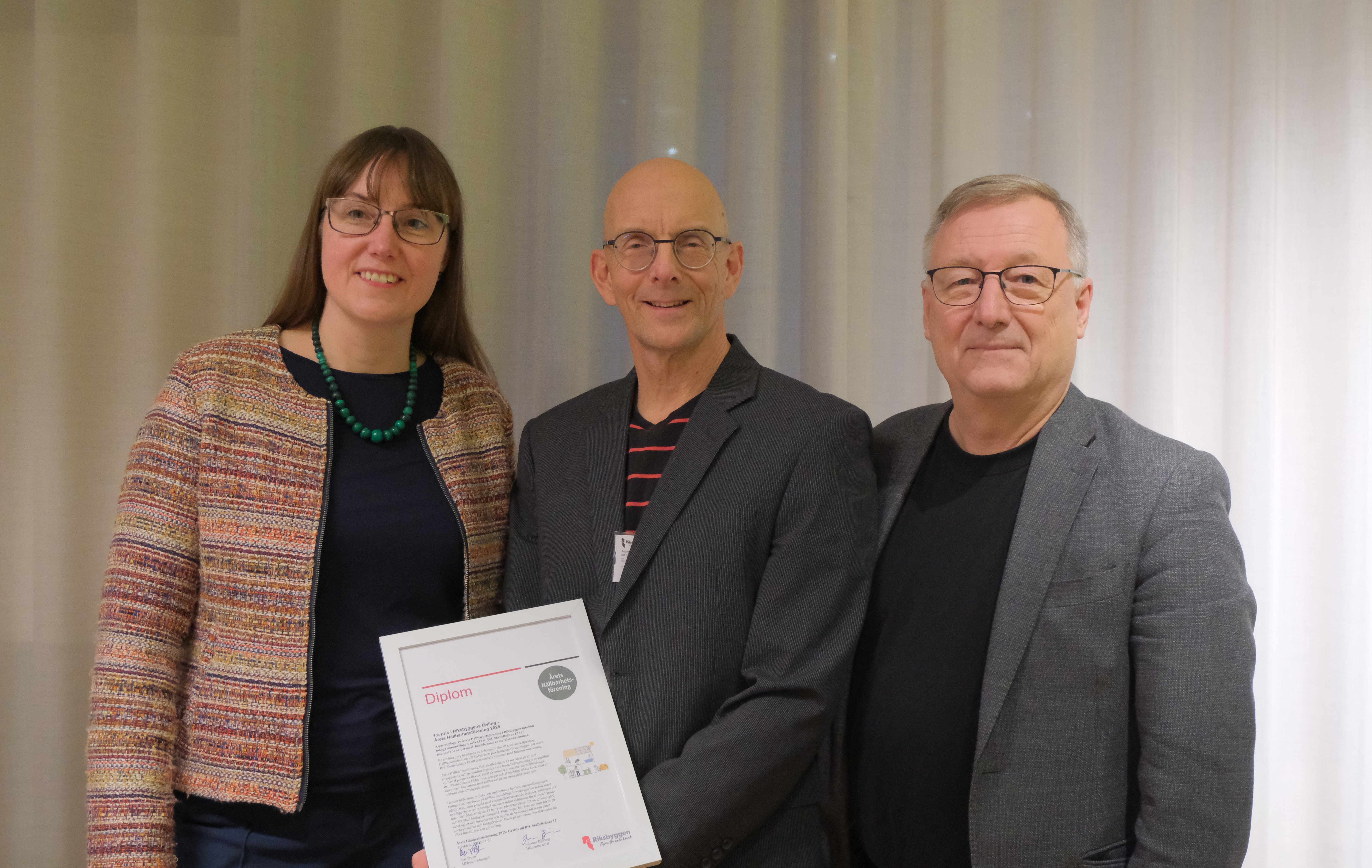 Tre personer står framför en gardin; en kvinna och två män, varav en håller ett diplom