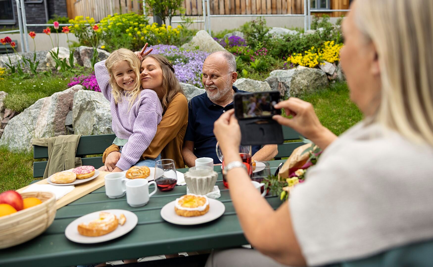 En grupp människor sitter vid ett utomhusbord med fika och mat, omgiven av en blomsterträdgård. En person fotograferar scenen med en mobiltelefon