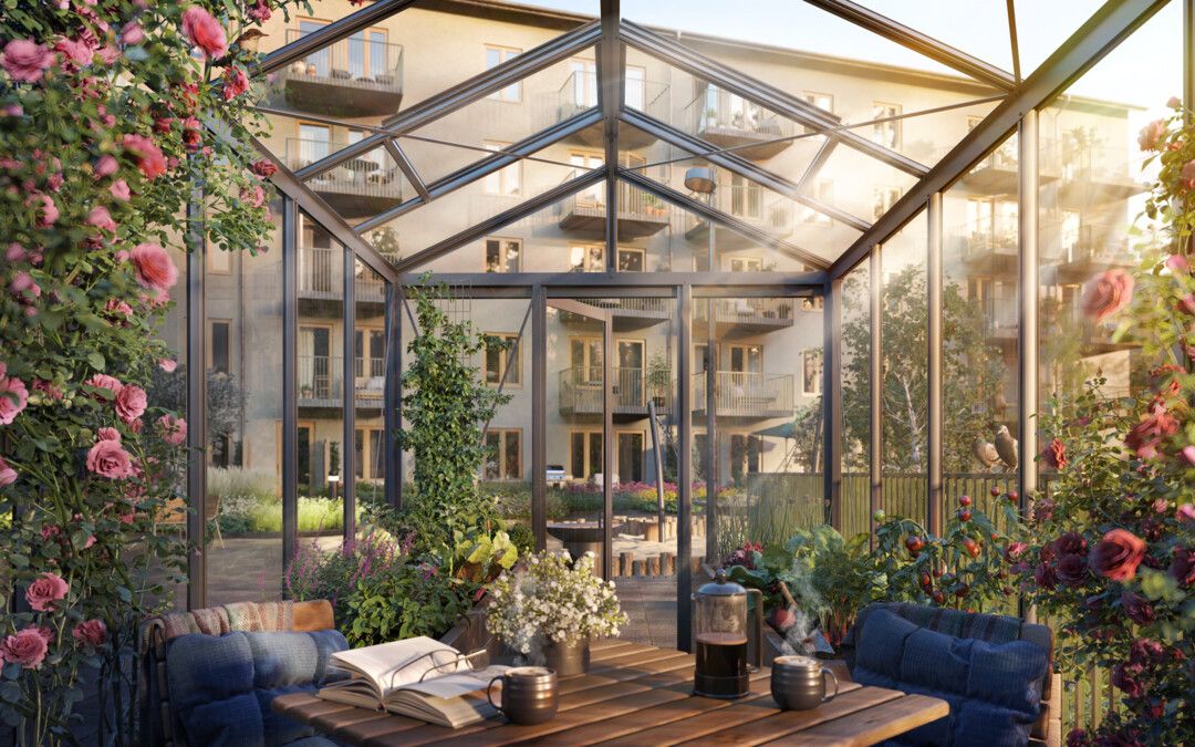 Växthus med glasväggar, omgiven av grönska och blommor, med utsikt över en innergård och bostadsbyggnader i bakgrunden. Bord med böcker, blommor och koppar är placerat inuti