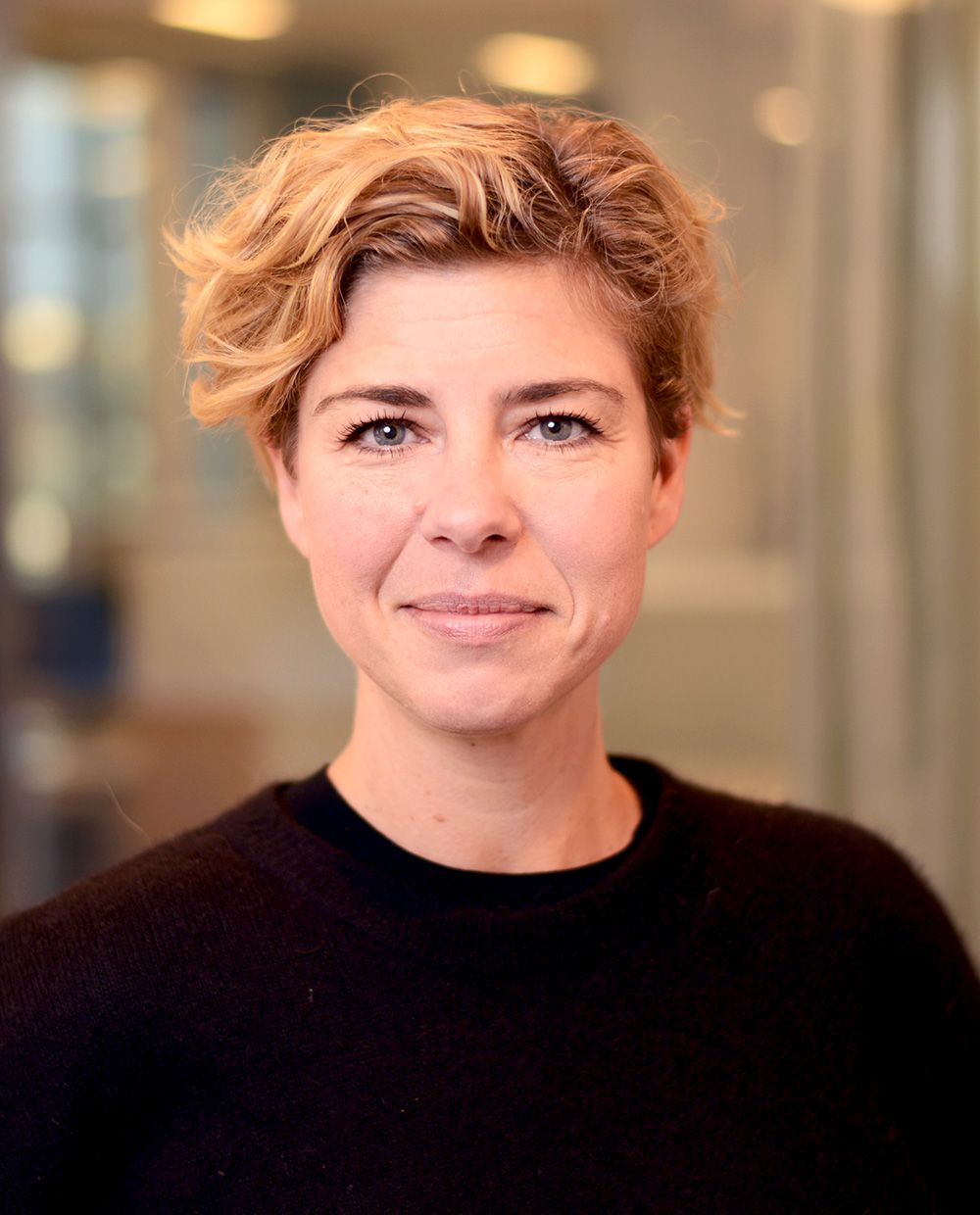 En person med kort, blont hår iklädd en svart tröja, stående framför en suddig bakgrund inomhus