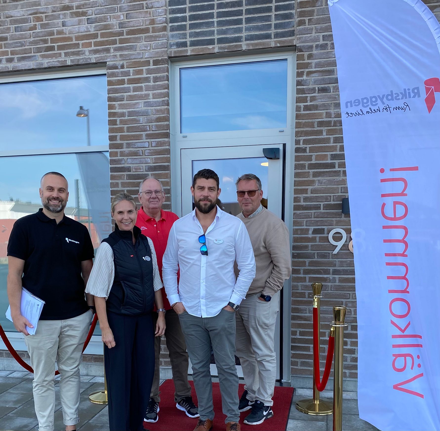 En grupp personer står utanför en byggnad med röd matta, repstolpar och en flagga med texten "Rikskyggen