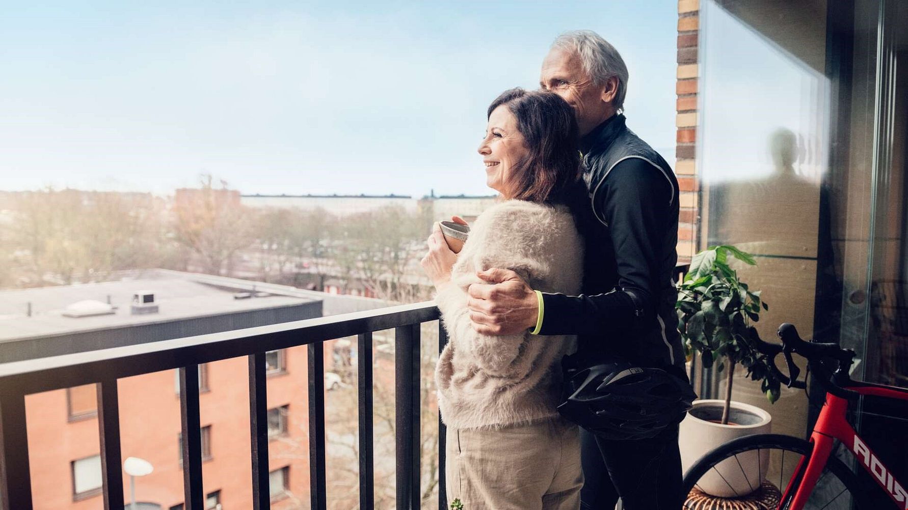 Två personer står på en balkong och tittar ut över ett stadslandskap, med ett cykelstyre synligt till höger och en krukväxt bredvid dem