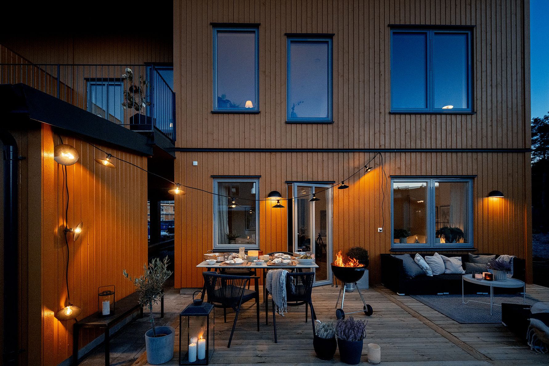 Modern träfasad med stor terrass på kvällen, inredd med matbord, stolar, soffgrupp, eldkorg och dekorativ belysning