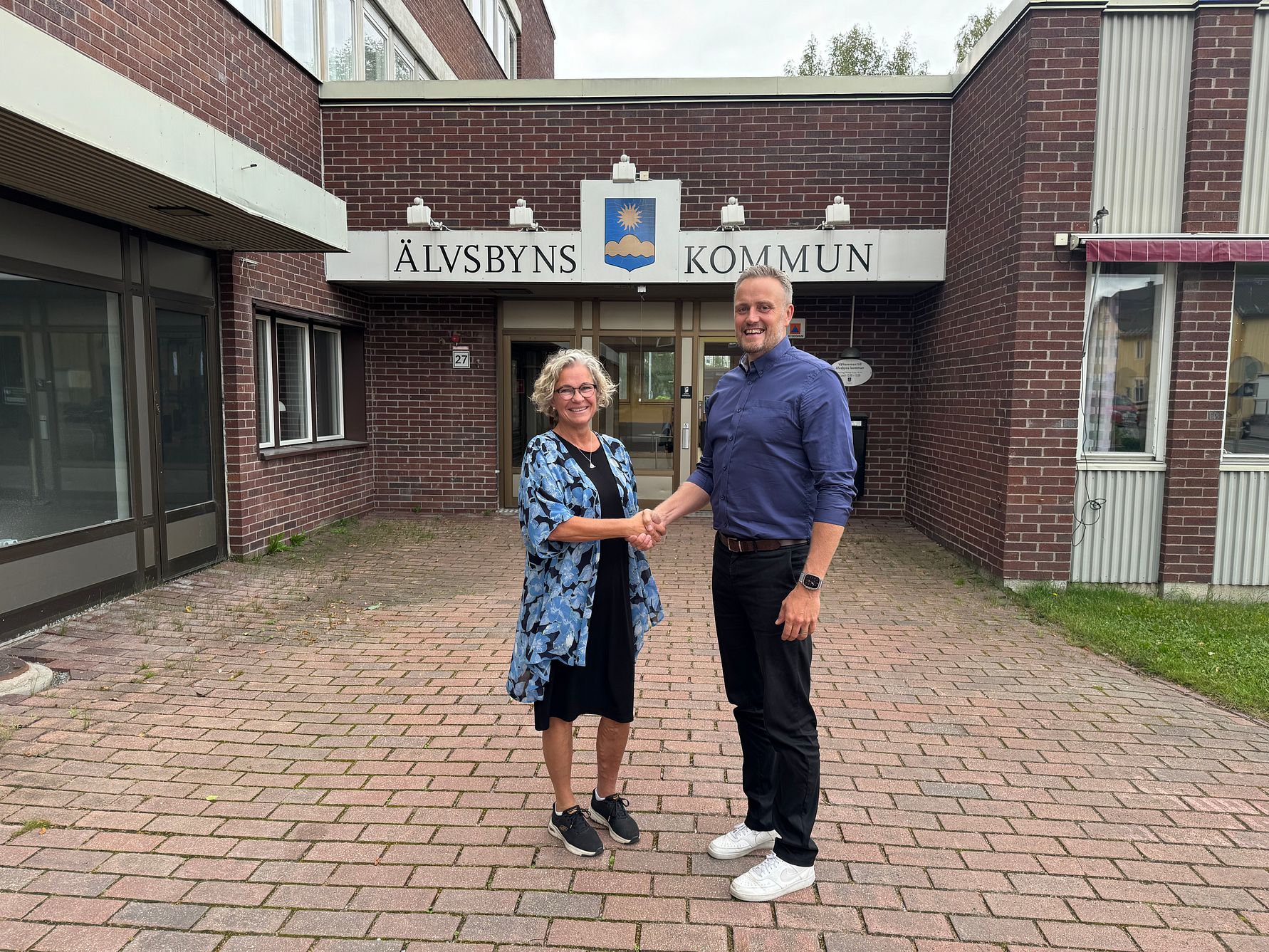 Två personer skakar hand framför byggnaden Älvsbyns kommun, som är gjord av tegel med en skylt ovanför entrén