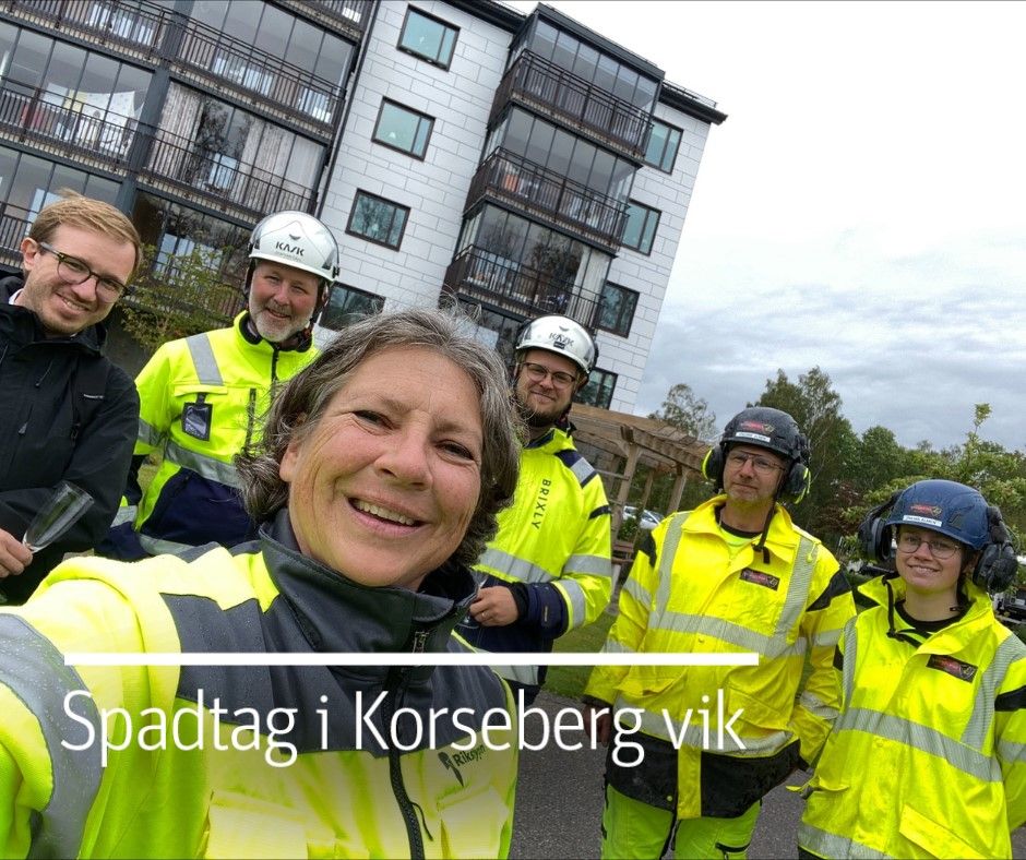 Personer i arbetskläder och hjälmar står framför en byggnad med vita fasader och balkonger. Texten "Spadtag i Korseberg vik" visas på bilden
