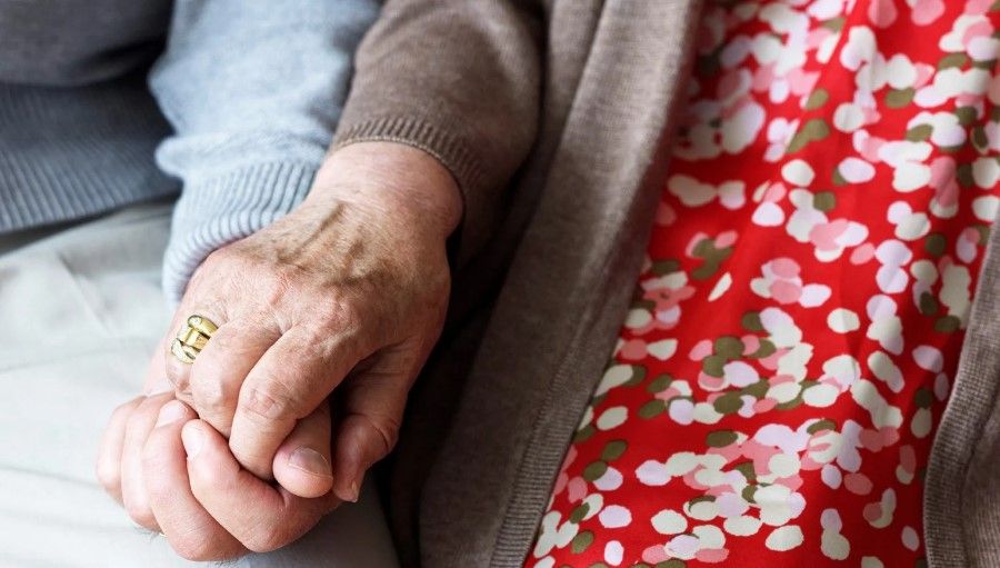 Närbild av äldre personer som håller varandra i handen, med en färgglad klänning och en brun kofta synlig
