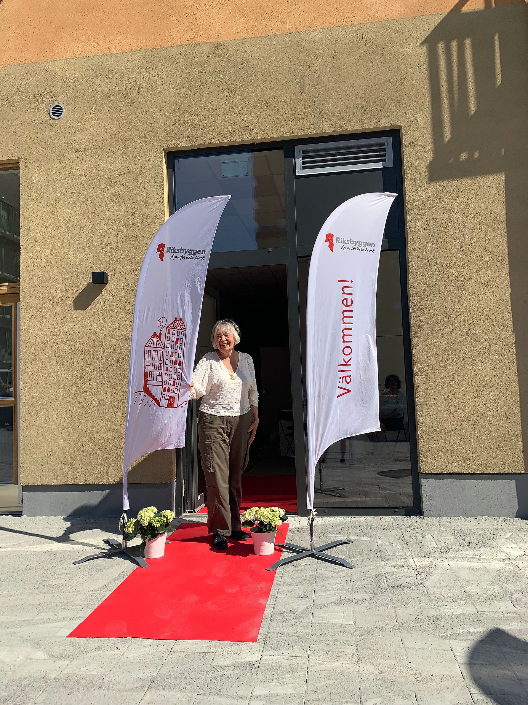 En person står på en röd matta framför en byggnad, flankerad av två vita flaggor med text och logotyp för Riksbyggen