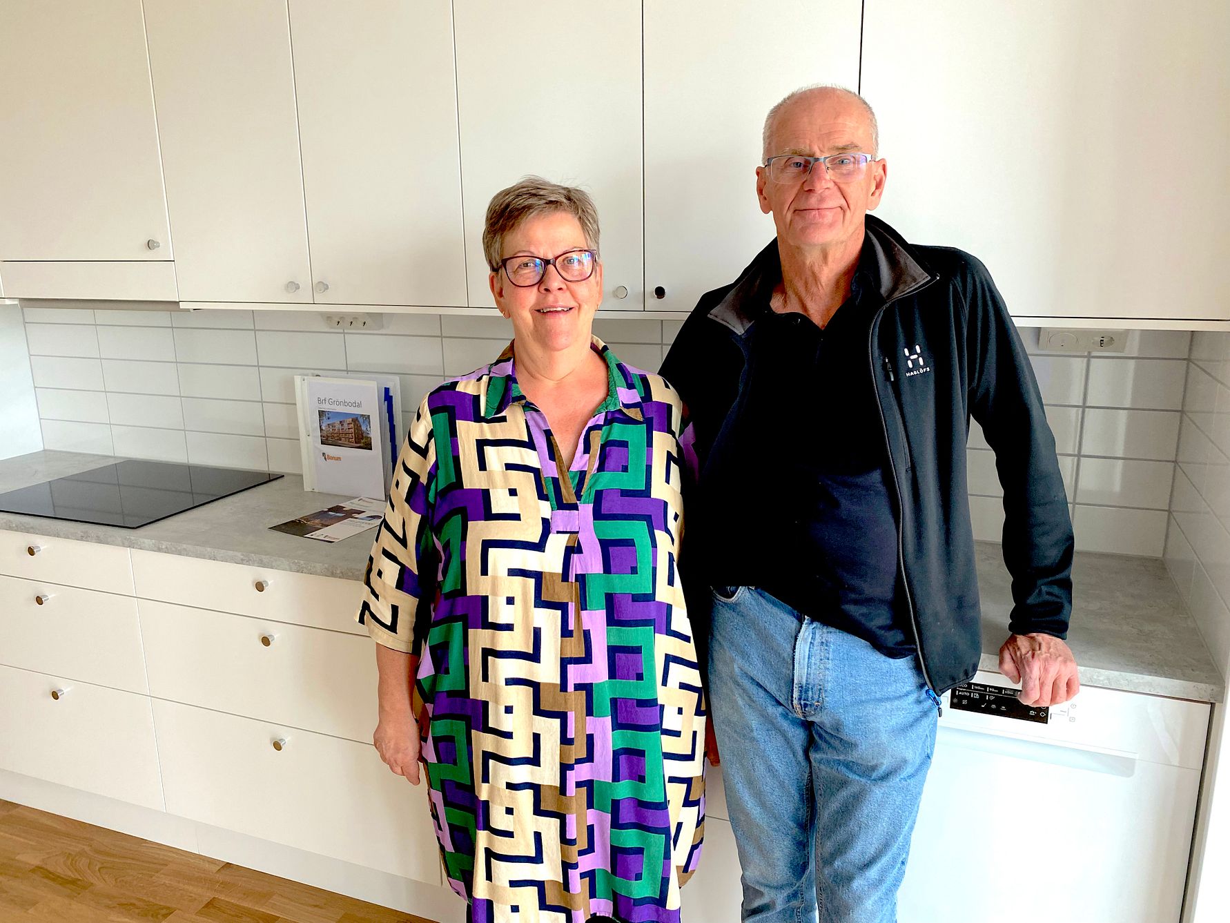 Två personer står i ett modernt kök med vita skåp, kaklade vita väggar och ljus bänkskiva