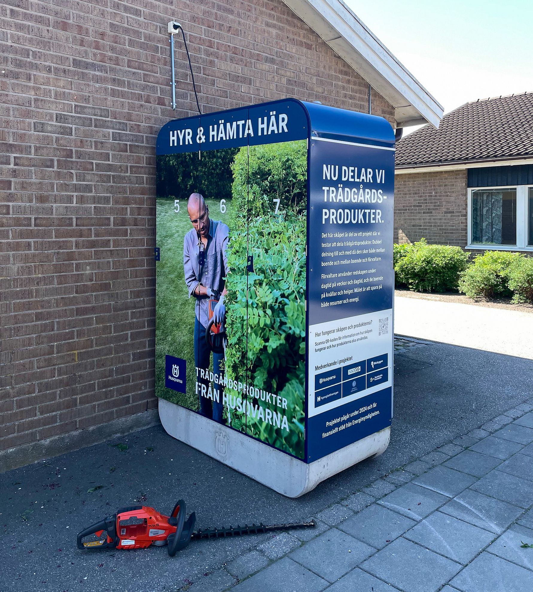 Hyra- och lånestation för trädgårdsprodukter från Husqvarna placerad utomhus vid en tegelvägg, med en beskärningsmaskin på marken bredvid