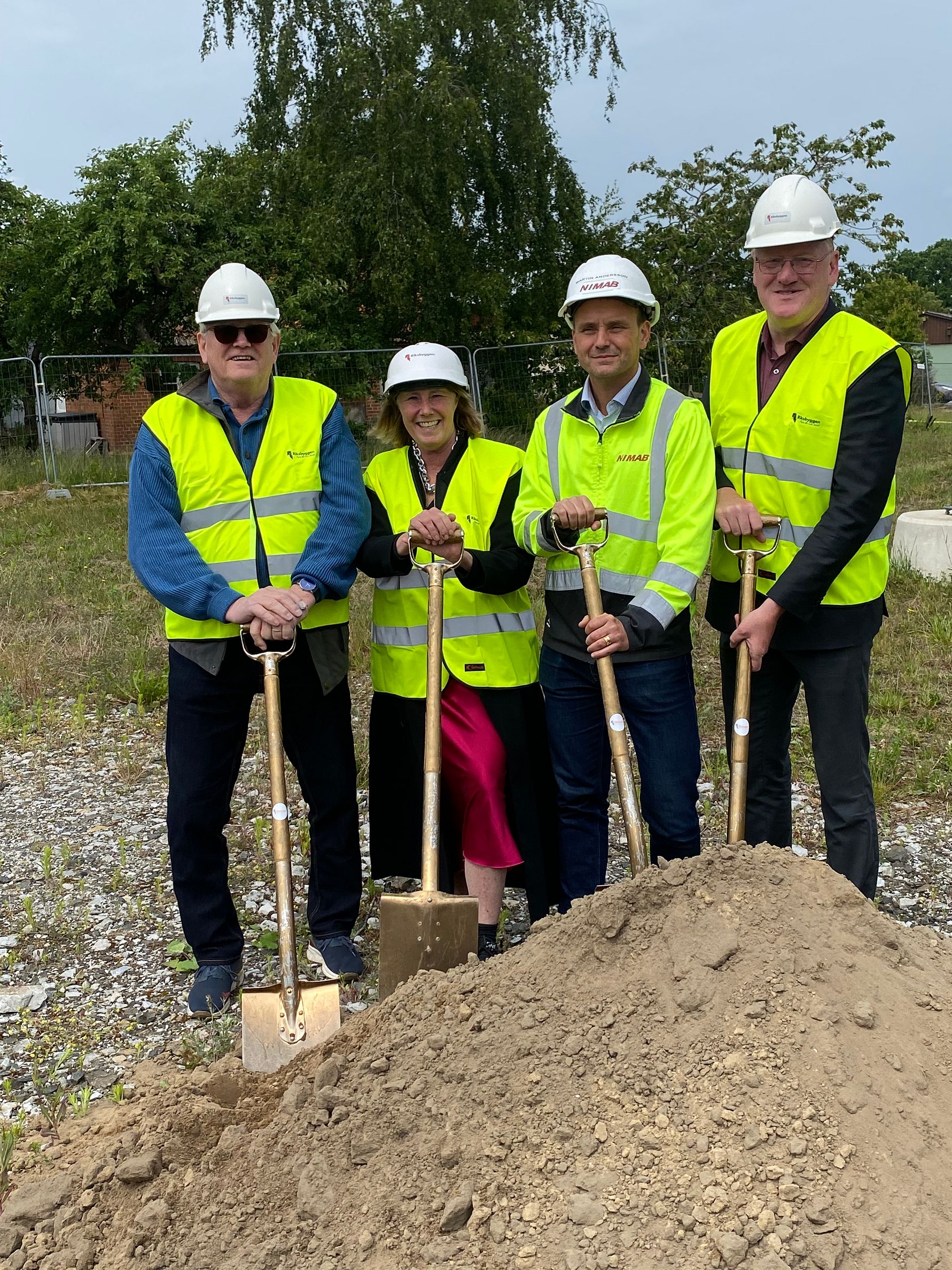 Fyra personer i reflexvästar och hjälmar står utomhus med spadar framför en hög med jord på en byggarbetsplats