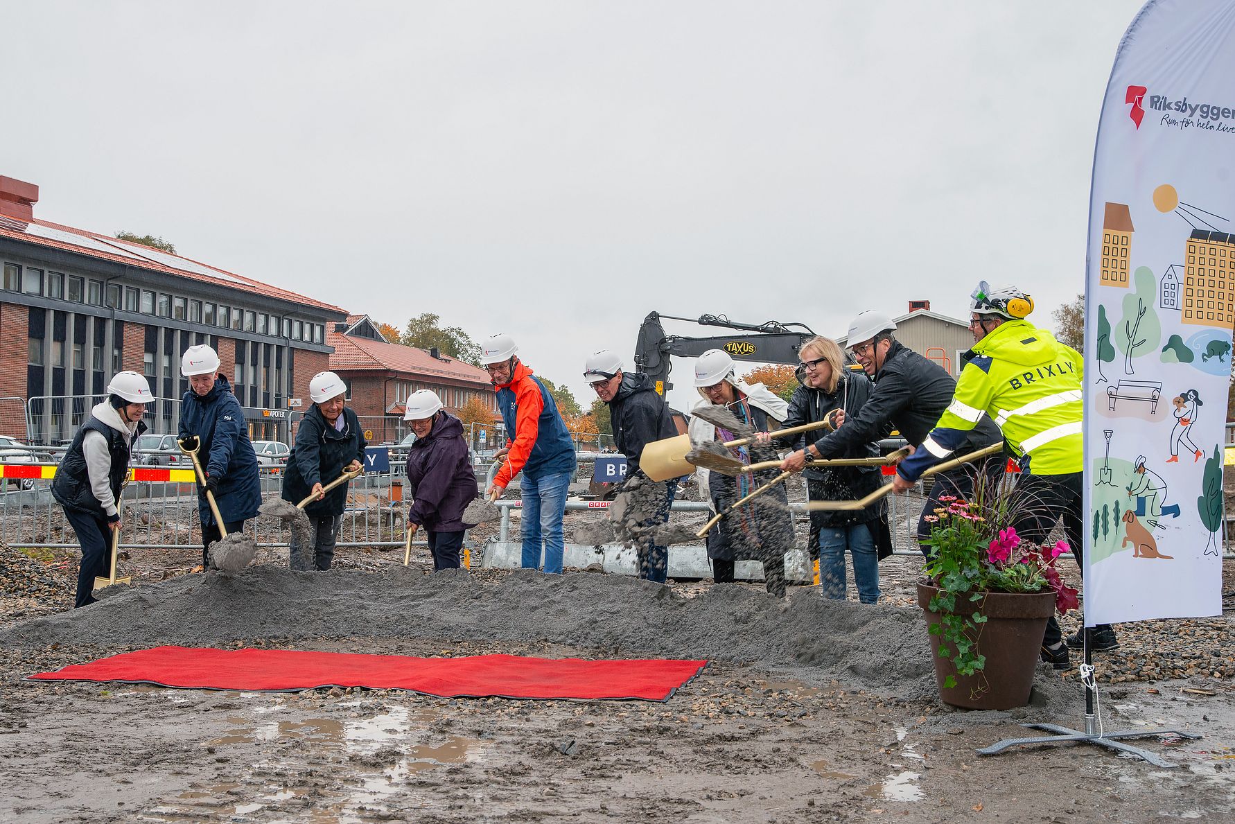 Grupp av människor med skyddshjälmar och reflexkläder deltar i en ceremoniell första spadtag vid en byggarbetsplats, med en grävmaskin och byggnader i bakgrunden