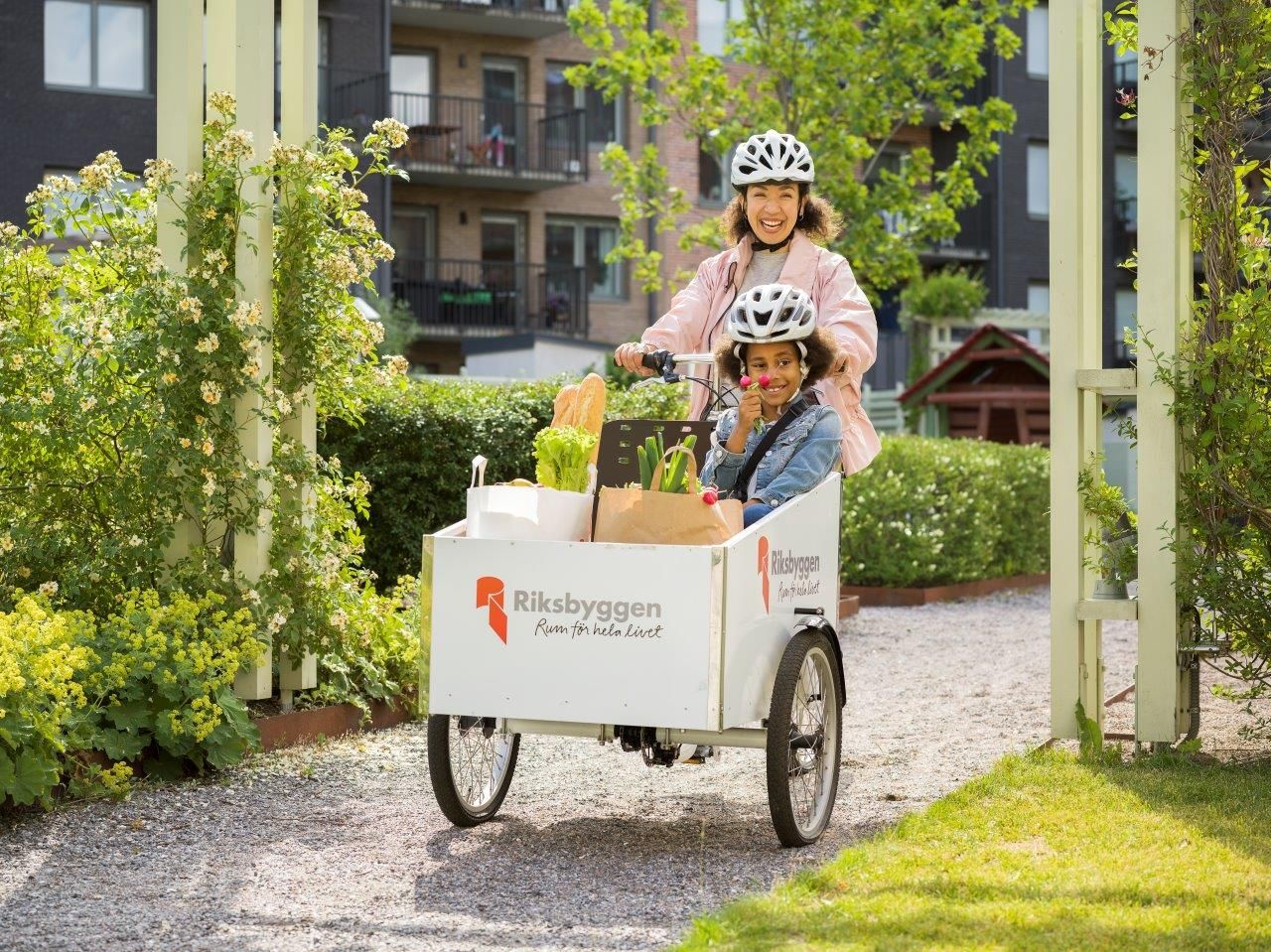 Två personer cyklar en lastcykel med Riksbyggen-logotypen, fylld med matvaror och en växt, på en grusad stig omgiven av grönska och bostadshus i bakgrunden