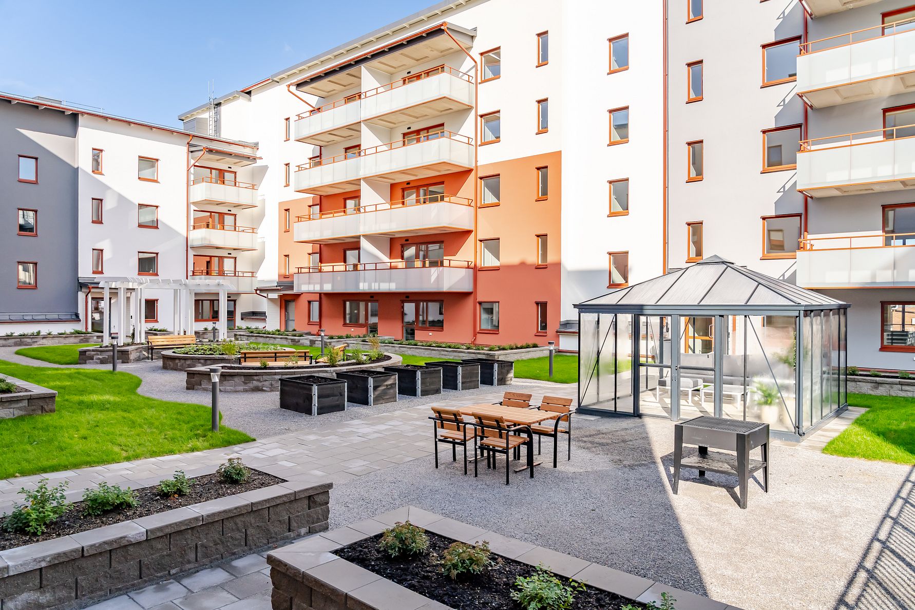 Bostadsgård med modern flerfamiljsbyggnad, grönskande gräsmattor, planteringslådor, sittmöbler och ett växthus under klarblå himmel