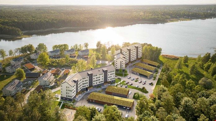Flygfoto över ett bostadsområde med flerfamiljshus, småhus och parkeringsplatser, beläget vid en sjö omgiven av skog och grönska