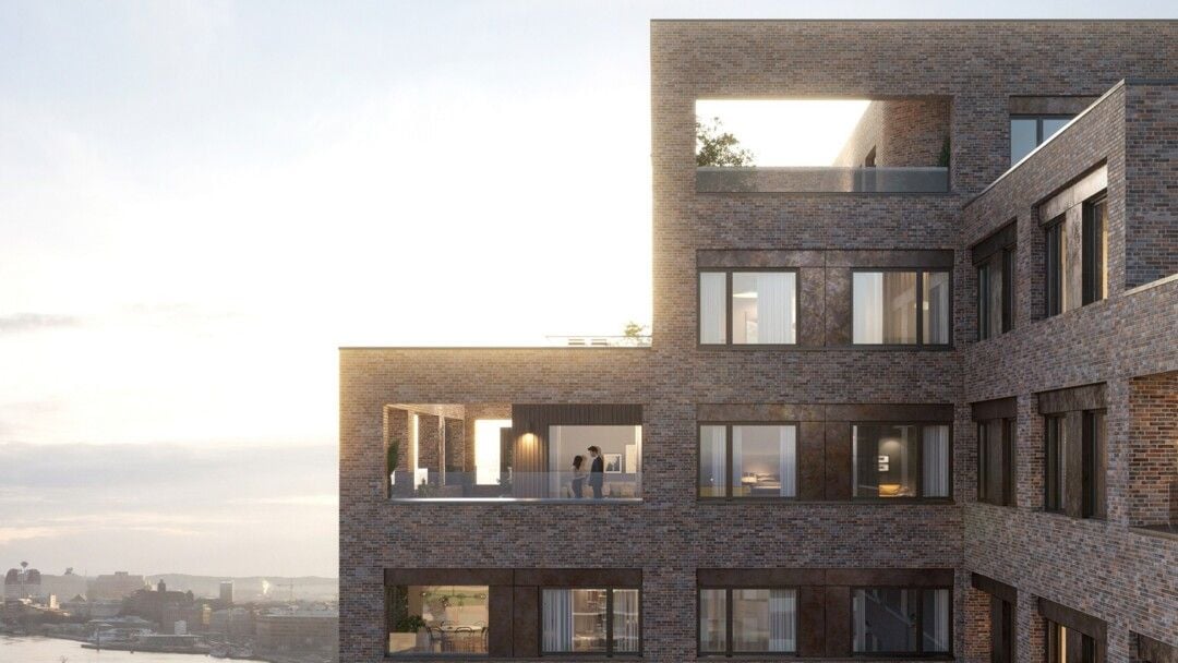 Tegelbyggnad med moderna lägenheter, balkonger och stora fönster under en ljus himmel med stadsvy i bakgrunden