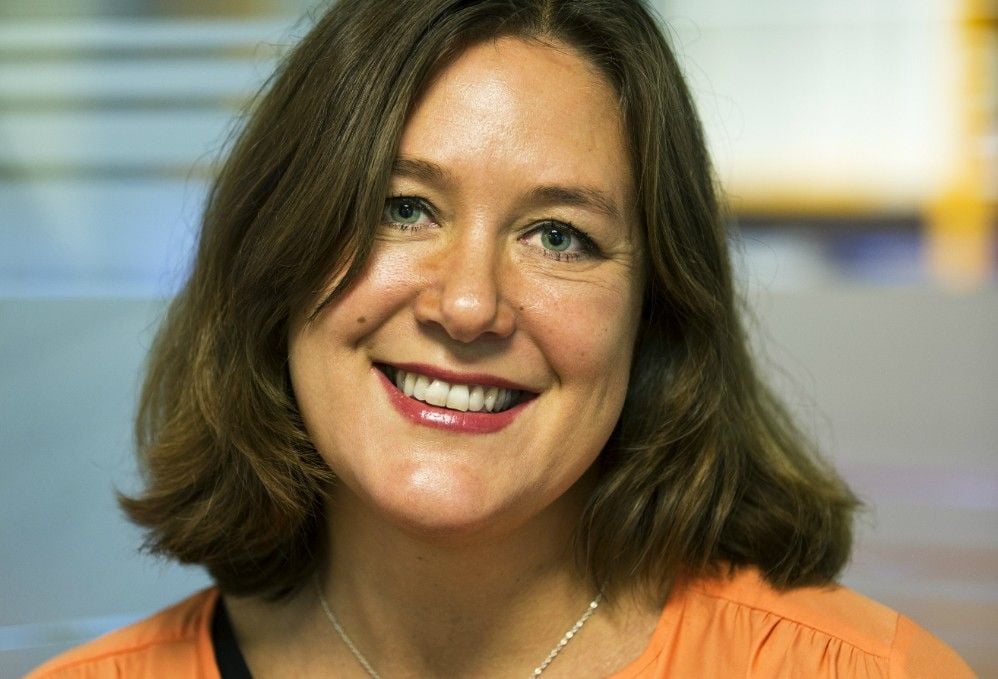 Kvinna med axellångt brunt hår, iklädd orange tröja och ett halsband, med en oskarp bakgrund i inomhusmiljö