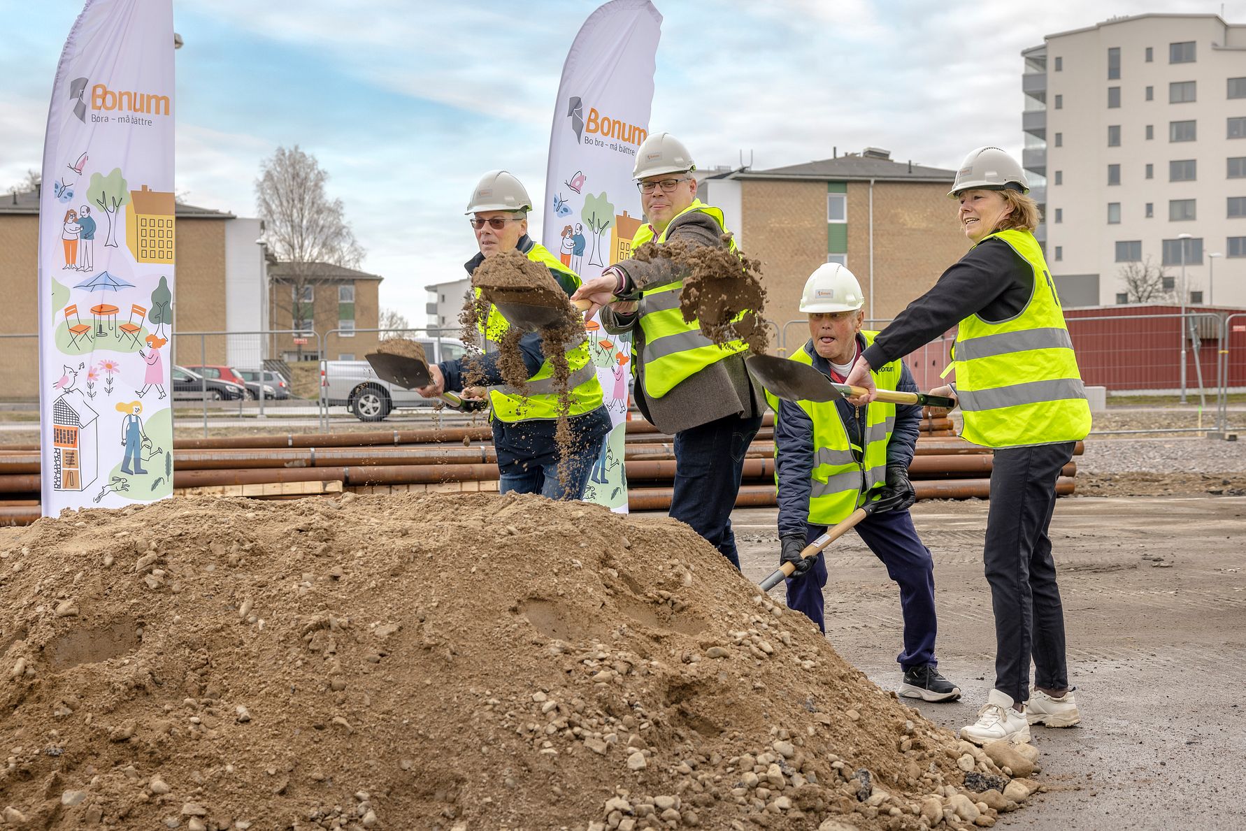 Flera personer i reflexvästar och hjälmar deltar i en symbolisk första grävning vid en byggarbetsplats, med byggnader och flaggor i bakgrunden