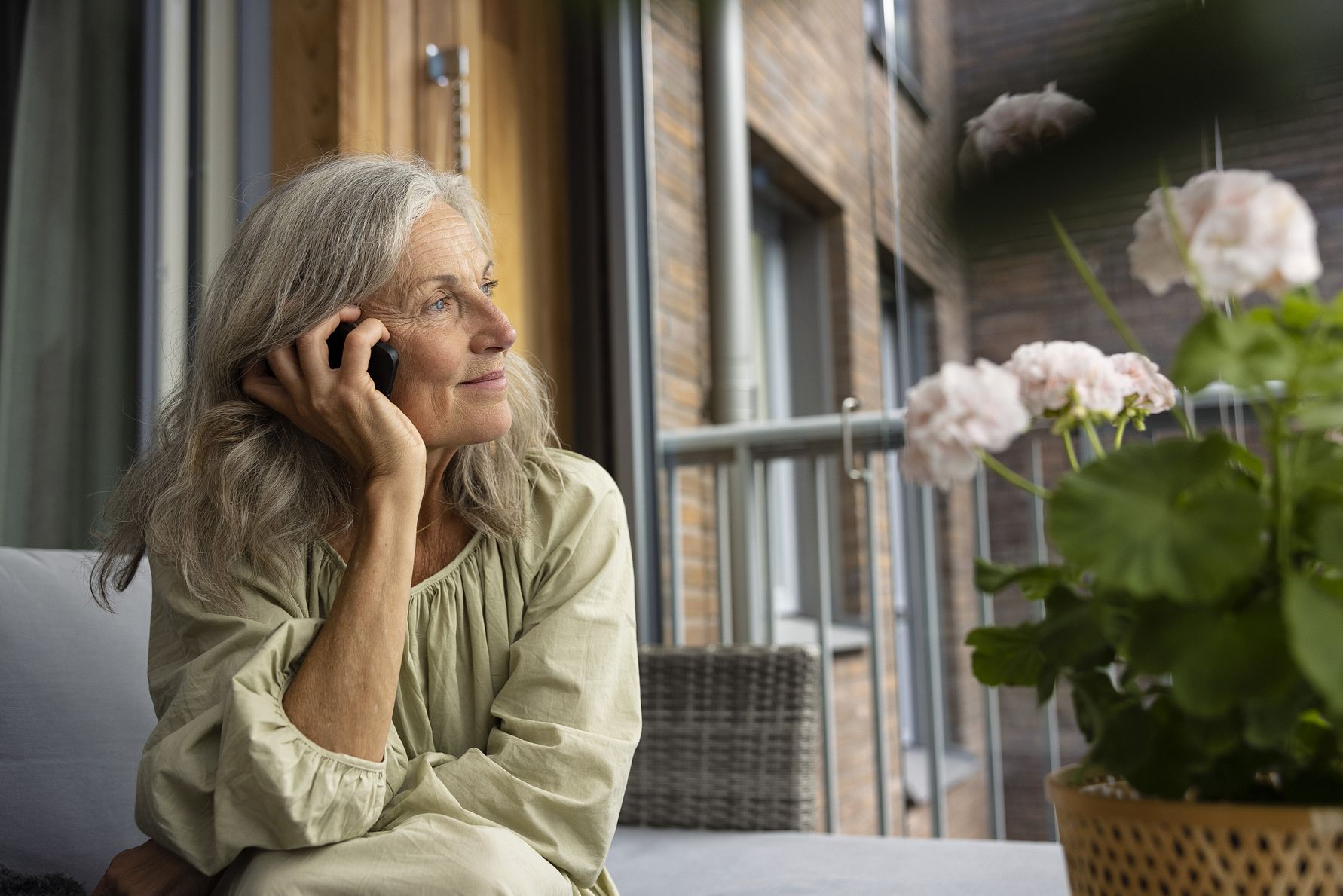 En person med grått hår sitter på en soffa utomhus och pratar i telefon, med blommor och en balkong i bakgrunden