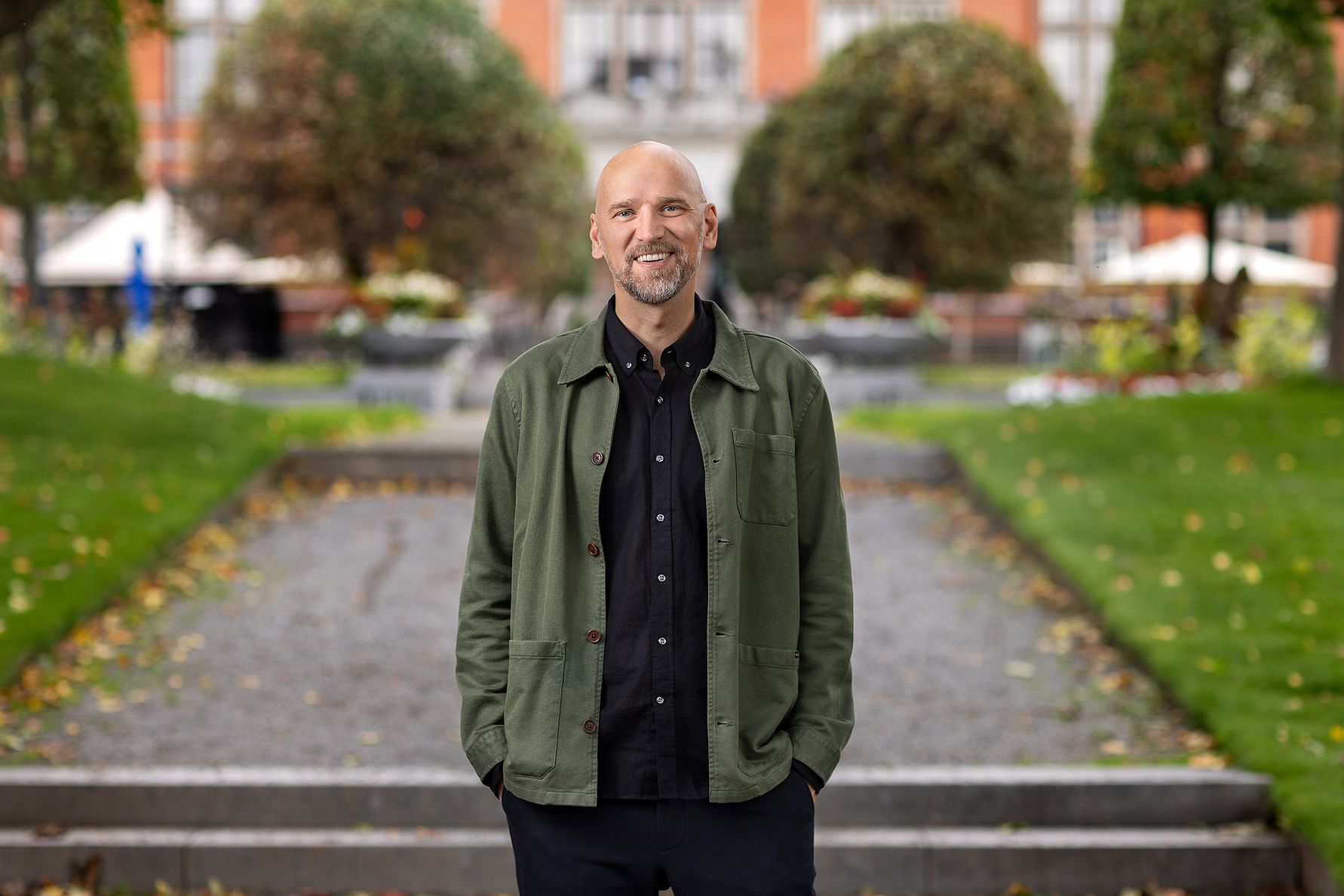 En person i en grön jacka och svart skjorta står på en grusgång i en park med gräs, buskar och träd i bakgrunden