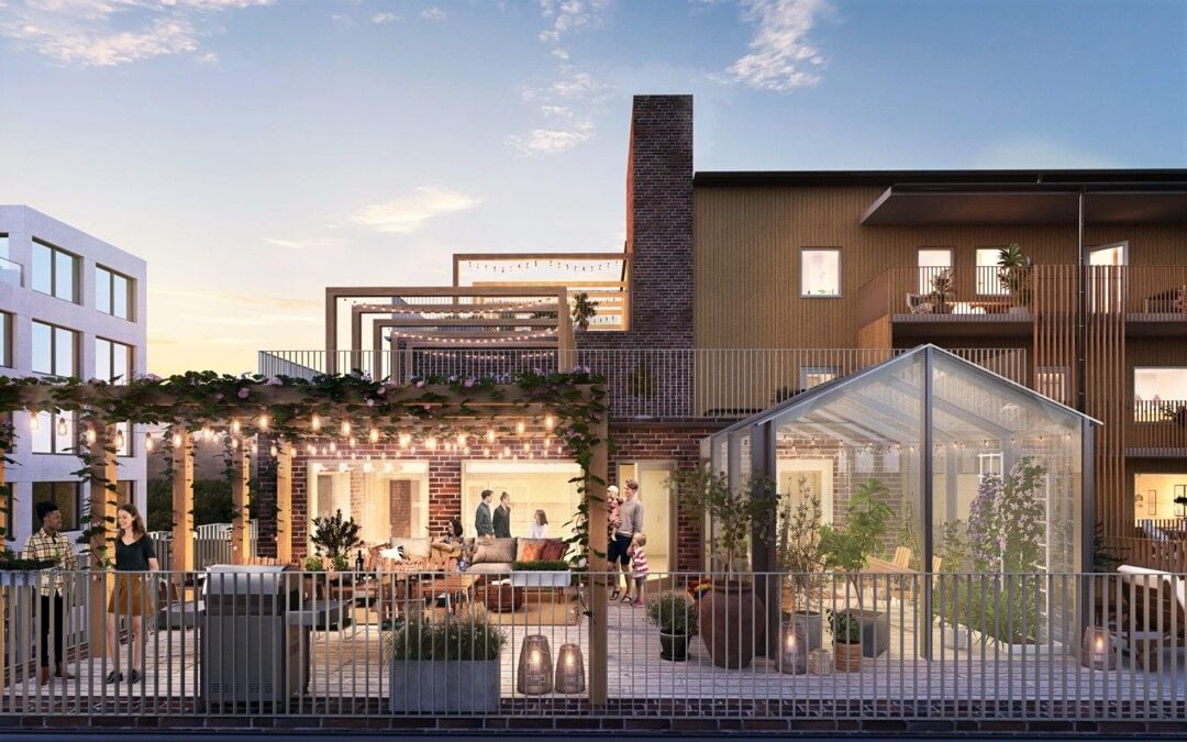 Modern bostadsområde med en rymlig terrass, växthus och utomhusbelysning, omgiven av grönska och byggnader i bakgrunden under en klar kvällshimmel