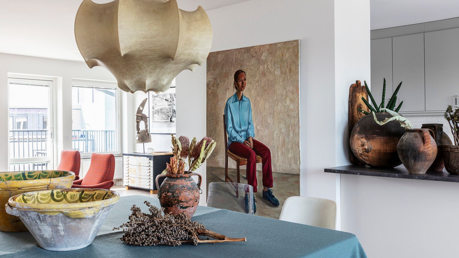Ett modernt matsalsområde med en turkos duk på bordet, omgiven av olika keramikföremål och torkade växter, med en stor dekorativ lampa ovanför samt ett porträtt och ett kök i bakgrunden