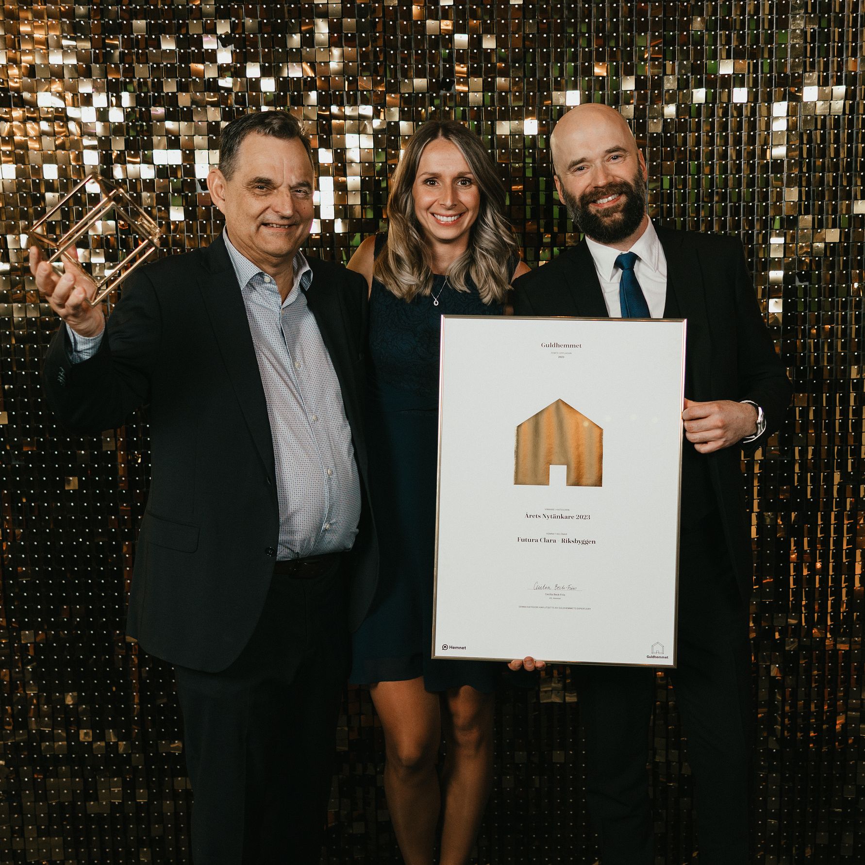 Tre personer klädda i formella kläder står framför en glittrande guldig bakgrund. En av dem håller ett pris, och en annan håller ett diplom med text