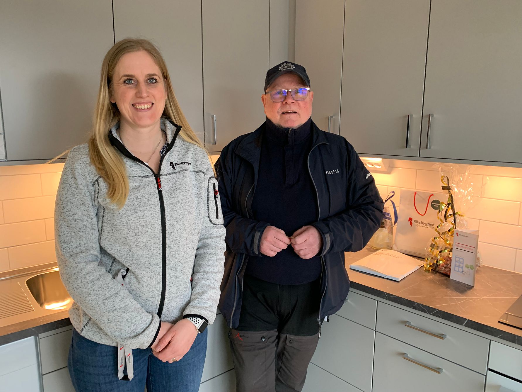 Två personer står i ett kök med grå skåpluckor och bänkskiva. Bakom dem syns ett paket och en dekorerad gåva på bänken