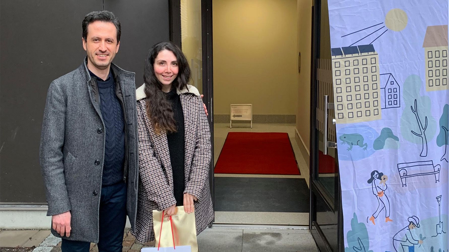 Två personer står framför en ingång med röd matta. I förgrunden syns en färgglad banner med stadsmotiv