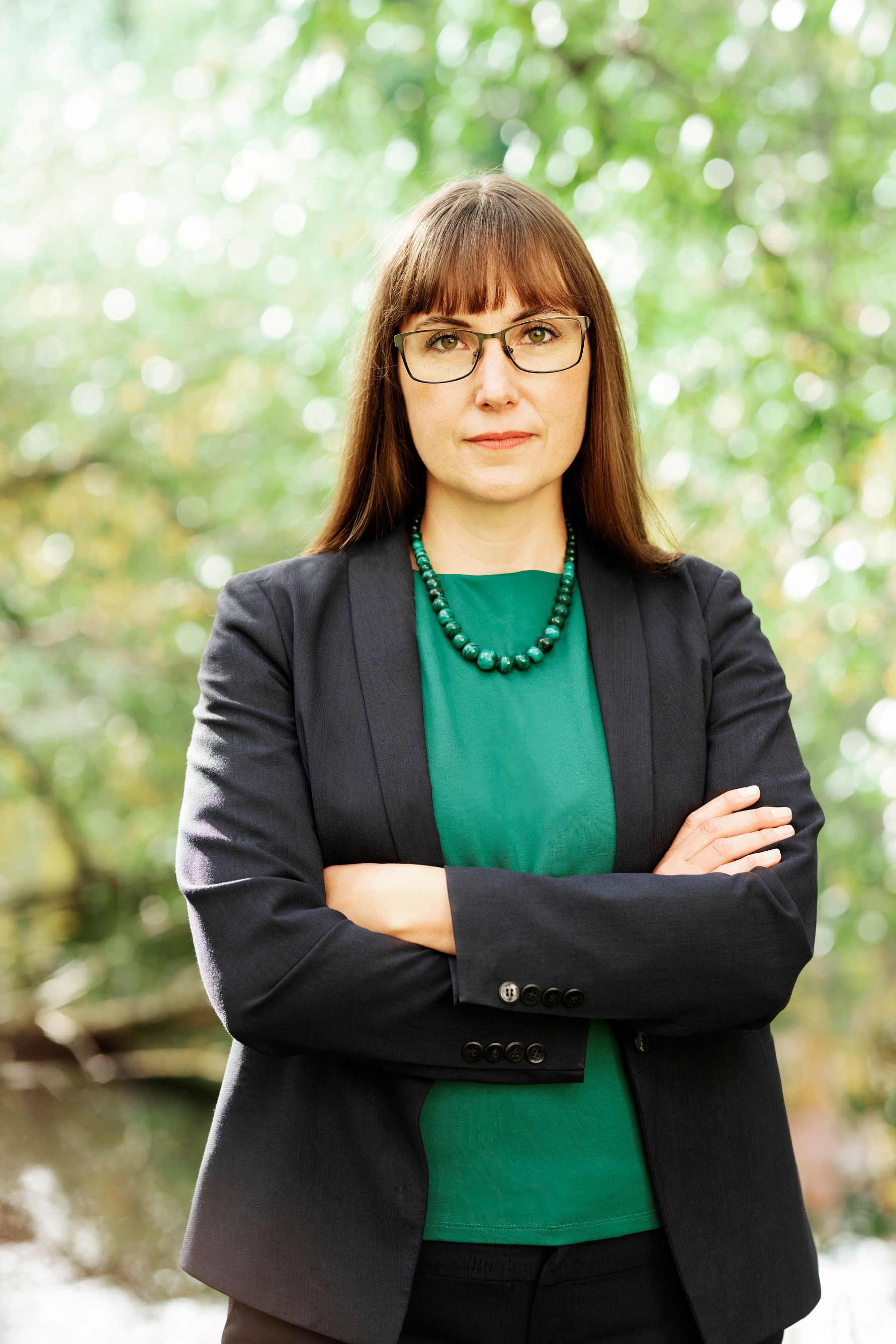Kvinna i mörk kavaj och grönt linne med korslagda armar stående framför grönskande bakgrund