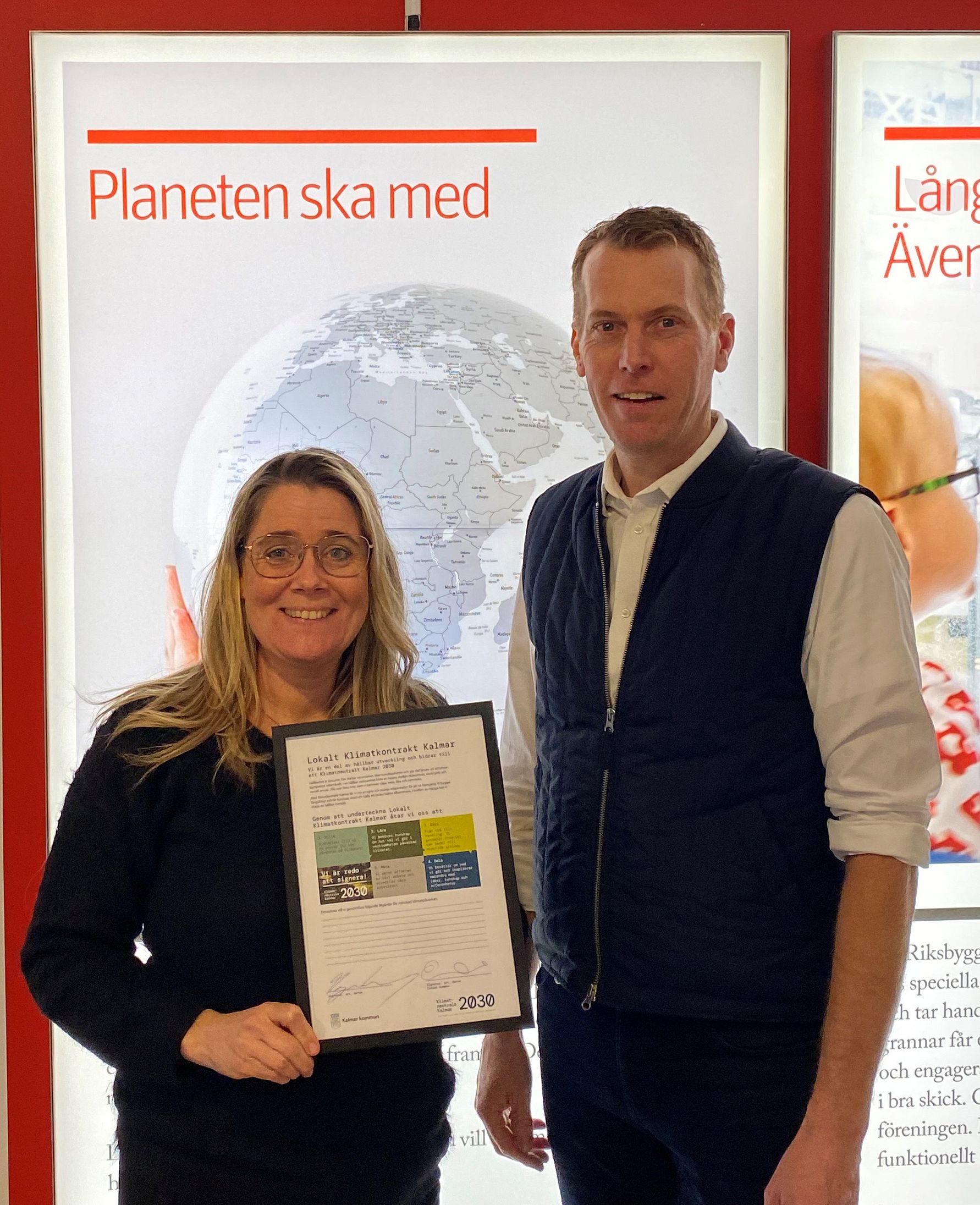 Två personer står framför en skylt med text och en bild av en jordglob. Personen till vänster håller ett dokument