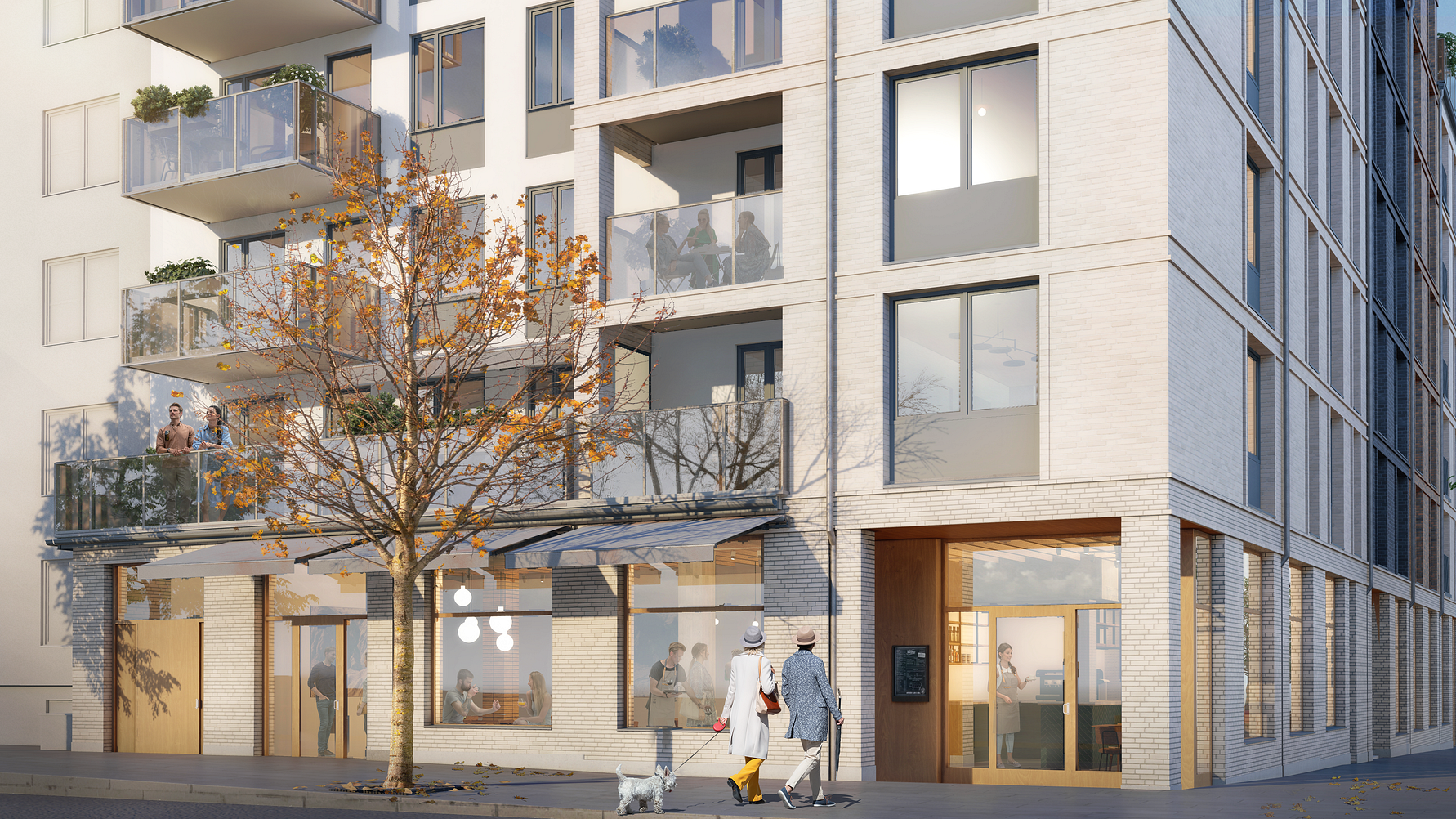 Flerbostadshus i modern arkitektur med stora fönster, balkonger och ett träd i höstfärger framför byggnaden. Två personer promenerar med en hund nära entrén