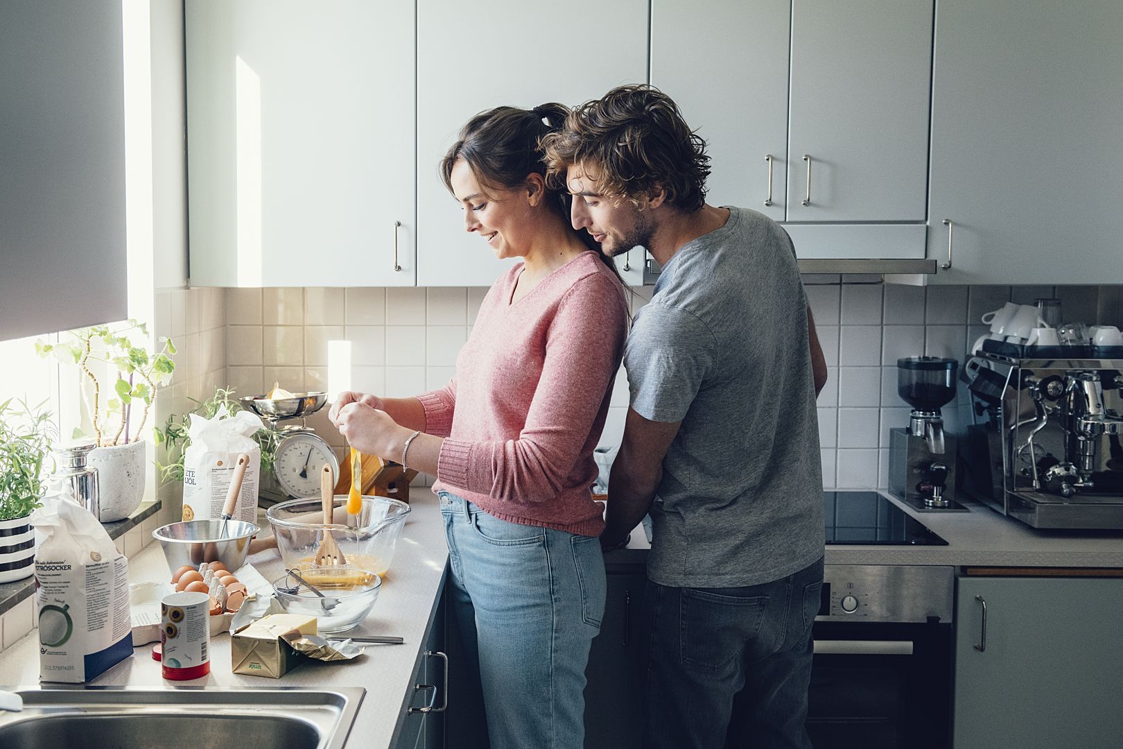 Två personer står i ett modernt kök och förbereder ingredienser, inklusive ägg, mjöl och smör, på en bänk