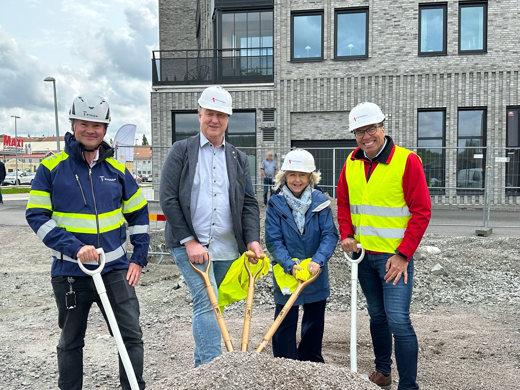 Fyra personer i bygghjälmar och reflexvästar håller i spadar vid en byggarbetsplats framför en nybyggd tegelfastighet. En grushög syns framför dem