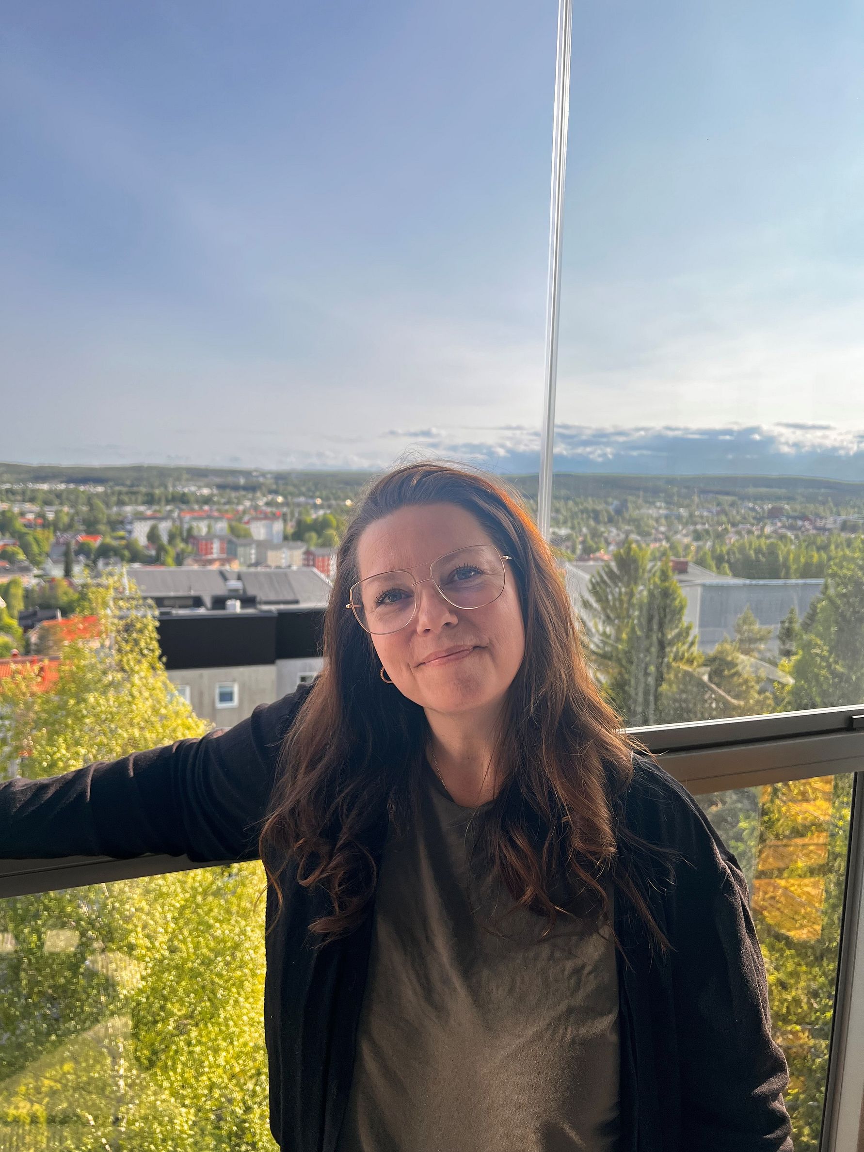 Person som står på en balkong med utsikt över en stad omgiven av träd och hus, med en klar himmel i bakgrunden