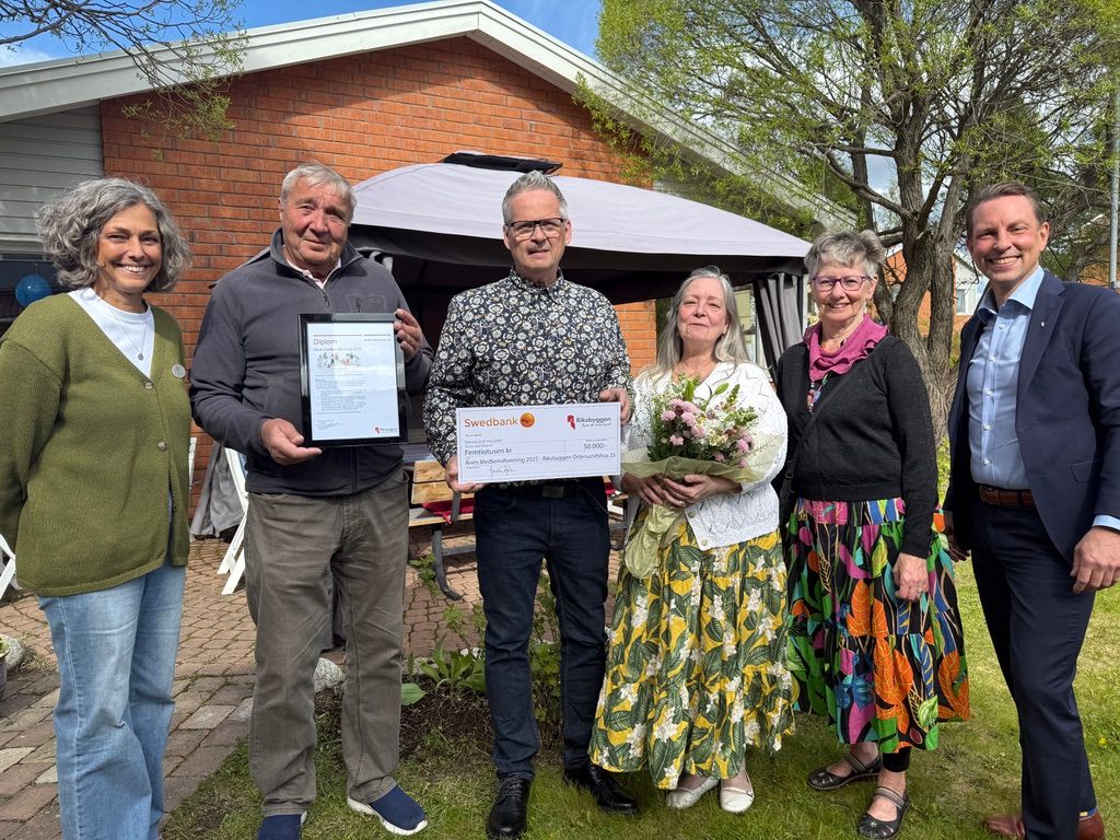 En grupp människor står utomhus framför ett tegelhus, där en person håller ett inramat dokument och en annan håller en stor check. En person har en bukett blommor i händerna